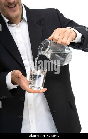 Junger, hübscher, hoher, schlanker weißer Mann, der Wasser aus einem Glaskrug in schwarzem Blazer gießt, der auf weißem Hintergrund isoliert ist Stockfoto