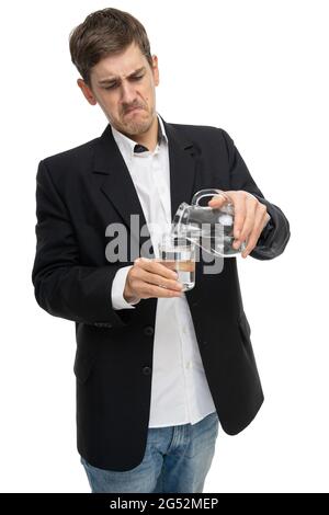 Junger, hübscher, großer, schlanker weißer Mann mit braunem Haar, der Wasser aus dem Glaskrug in ein kleines Glas in schwarzem Blazer auf weißem Hintergrund eingießt Stockfoto