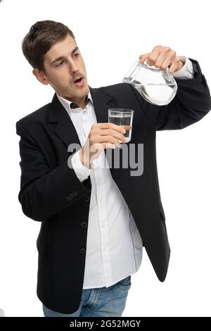 Junger, hübscher, großer, schlanker weißer Mann mit braunem Haar, der Wasser aus dem Glaskrug in ein kleines Glas in schwarzem Blazer auf weißem Hintergrund eingießt Stockfoto