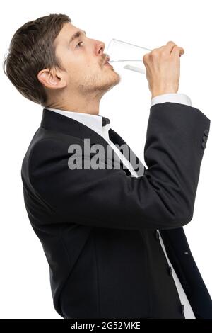 Junger, hübscher, großer, schlanker weißer Mann mit braunem Haar, der einen Schluck aus einem Glas Wasser in schwarzem Blazer nimmt, der auf weißem Hintergrund isoliert ist Stockfoto