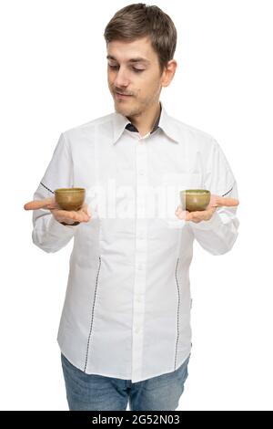 Junger, hübscher, großer, schlanker weißer Mann mit braunem Haar, der zwei Teetassen in weißem Hemd auf weißem Hintergrund hält Stockfoto