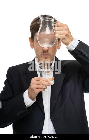 Junger, hübscher, großer, schlanker weißer Mann mit braunem Haar, der durch einen Wasserkrug aus Glas in schwarzem Blazer auf weißem Hintergrund isoliert schaut Stockfoto