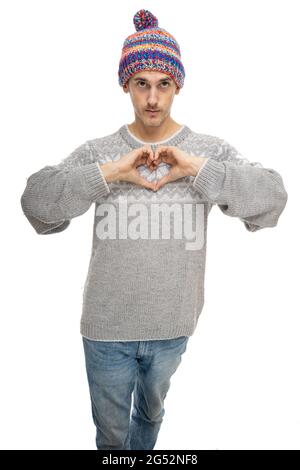 Junger, hübscher, großer, schlanker weißer Mann mit braunem Haar, der ein Herzzeichen in einem grauen Pullover mit farbenfrohem Winterhut auf weißem Hintergrund macht Stockfoto