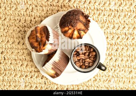 Drei duftende Schokoladenkuhmuffins mit einer Tasse Kaffee auf einem weißen Teller, Nahaufnahme, auf einer Strohmatte, Draufsicht. Stockfoto