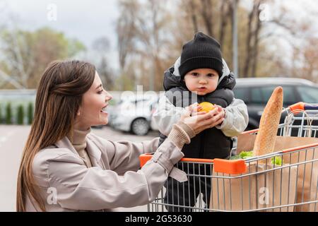 Glückliche Frau, die auf den kleinen Jungen schaut, der frische Orange hält, während er im Einkaufswagen sitzt Stockfoto