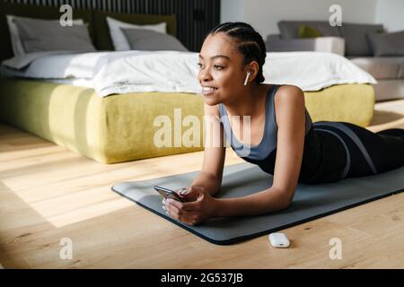 Attraktive junge lächelnde afrikanerin, die im Schlafzimmer mit Kopfhörern Musik hört und das Mobiltelefon benutzt Stockfoto
