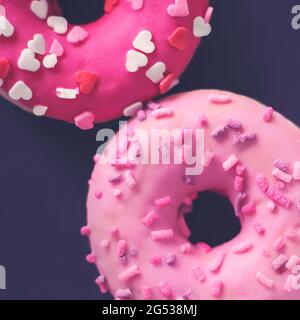 Draufsicht auf leckere süße rosa Donuts mit Streuseln in Form von Herzen, die auf einem dunkelvioletten Hintergrund liegen. Ein angenehmes und romantisches Geschenk für Valen Stockfoto