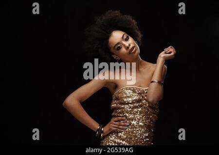 Nachdenkliche junge afrikanische Frau in Kleid, die über schwarzem Wandhintergrund steht und posiert Stockfoto