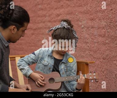 Junger hispanischer Mann lehrt lockiges Mädchen, Ukulele im Freien Musikkonzept zu spielen Stockfoto