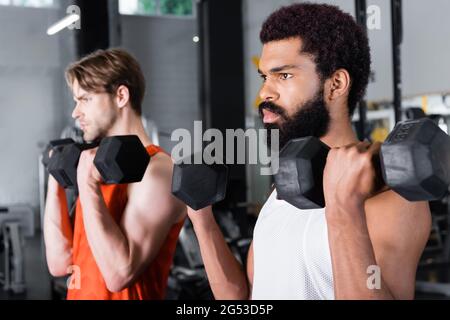 Bärtiger afroamerikanischer Mann, der mit Kurzhanteln trainiert, in der Nähe von verschwommenem Freund Stockfoto