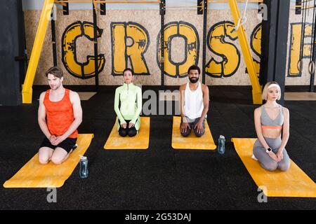 Interrassische Menschen, die auf Fitnessmatten im Blitzschlag sitzen, posieren in der Nähe von Sportflaschen Stockfoto