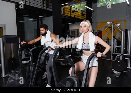 Blonde Frau und afroamerikanischer Mann trainieren im Fitnessstudio auf Heimtrainern Stockfoto