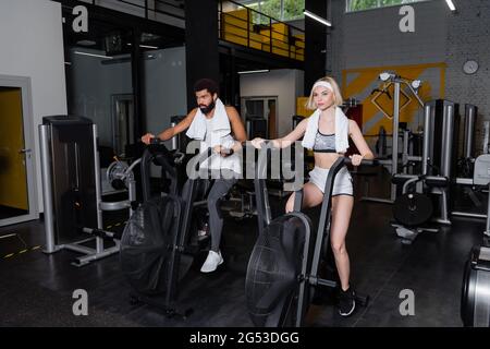 Blonde Frau und bärtiger afroamerikanischer Mann trainieren auf Trainingsgeräten im Fitnessstudio Stockfoto