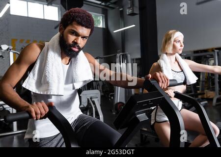 Bärtiger afroamerikanischer Mann, der an einem Trainingsgerät in der Nähe einer blonden Frau arbeitet Stockfoto