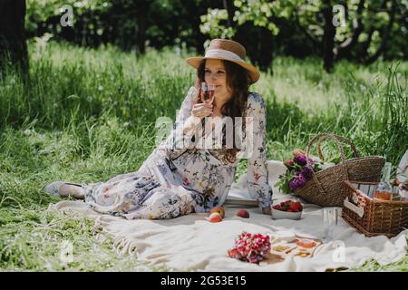 Junge Frau im Sommerkleid und Strohhut, die mit Rosenwein, Käse, frischem Obst und Baguette auf der Decke sitzt. Stockfoto