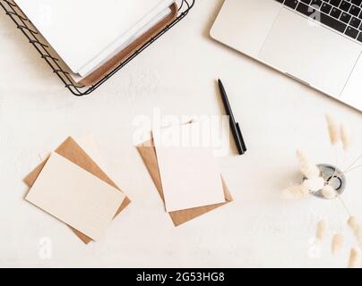 Schreibtisch mit Laptop, Aufbewahrungskorb, trockene Blumen in Vase, Umschläge und Postkarten auf weißem Hintergrund. Flach liegend, Draufsicht Stockfoto