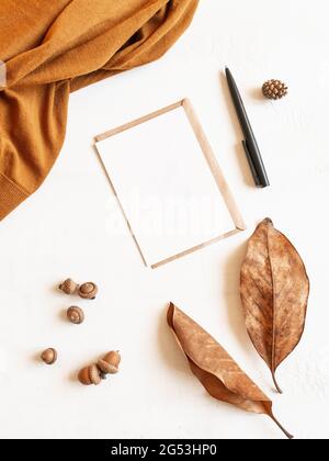 Rahmen aus braunem Pullover, Herbstblatt und Eicheln, Umschlag und Postkarte auf weißem Hintergrund. Draufsicht. Kopierbereich. Minimale Herbstzusammensetzung Stockfoto