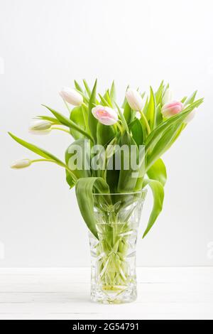 Blumenstrauß aus Frühlingsblumen in zartem Weiß und Rosa in einer transparenten Kristallvase auf weißem Holztisch Stockfoto