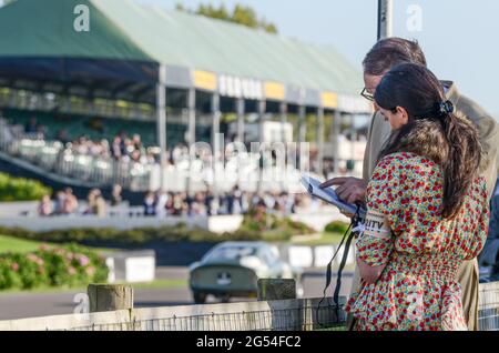 Männlich und weiblich in Vintage-Kostümen, die während des Rennens beim Goodwood Revival 2011 das Rennprogramm studieren. Tribüne und klassischer Rennwagen Stockfoto