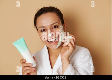 Close-up Beauty Portrait einer jungen Frau mit einem schönen toothy Lächeln Anwendung einer Gesichtscreme oder Peeling auf ihr Gesicht stehend isoliert über beige Backgr Stockfoto