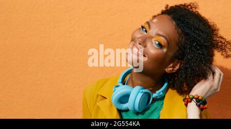 Porträt einer lachenden Frau mit Vitiligo in blauen Kopfhörern und bunten Kleidern auf dem Hintergrund der orangefarbenen Stadtmauer. Strahlender Lifestyle Stockfoto