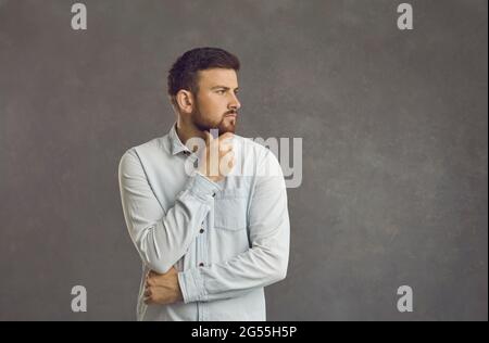 Ernster Mann, der auf grauem Hintergrund steht, denkt und blickt zur Seite auf den Kopierraum Stockfoto