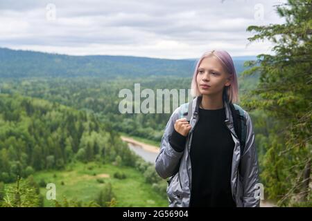 Das Mädchen im Teenageralter mit Rucksack geht auf dem Gipfel des Berges vor dem Hintergrund der bewaldeten Hügel und des Flusses spazieren Stockfoto