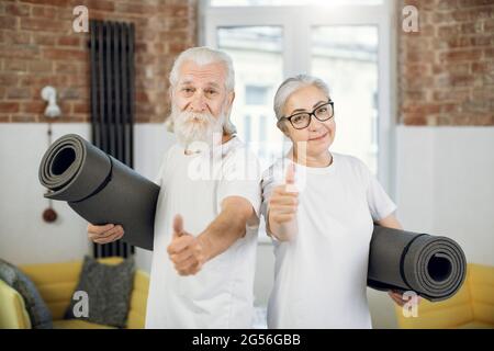 Mann und Frau im positiven Alter, die in Sportkleidung gekleidet sind, Yogamatte halten und den Daumen hoch zeigen, während sie im hellen Wohnzimmer stehen. Konzept von Menschen, aktiven Lebensstil und Ruhestand. Stockfoto