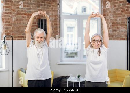 Angenehme reife Frau und Mann in Sportkleidung stehen zu Hause mit geballten Händen über dem Kopf. Aktives Paar, das morgens Übungen macht. Stockfoto