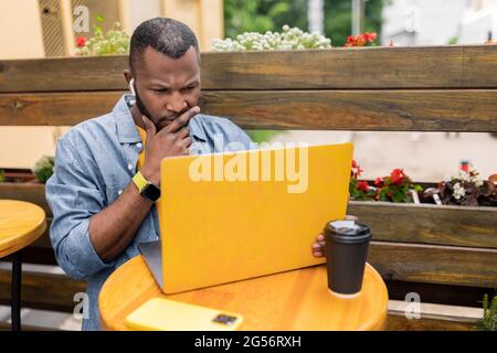 Konzentrierter afroamerikanischer Geschäftsmann, der im Café mit Laptop in kabellosen Kopfhörern arbeitet, qualifizierter junger freiberuflicher männlicher CEO, Geschäftsmann Stockfoto