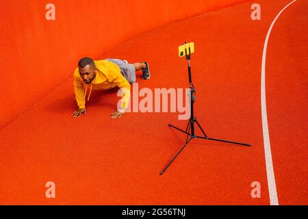 Afroamerikanischer sportlicher Mann schaut lächelnd auf die Telefonkamera, während er Plank-Übungen macht und mit der Webcam auf dem Smartphone auf dem Stativ das Training trainiert. Positiv Stockfoto