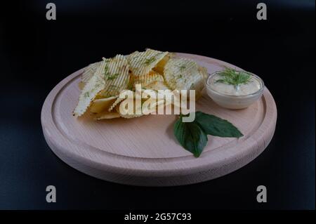 Kartoffelchips mit Dill und Sauce auf einem runden Holzbrett. Isolierter schwarzer Hintergrund. Stockfoto