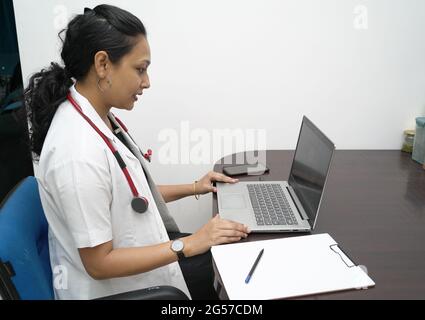 Eine südindische Ärztin in den 30er Jahren mit Laptop und Verordnungsplatine mit weißem Mantel und rotem Stethoskop auf weißem Hintergrund.Online-medizinische Beratung Stockfoto