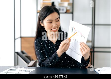 Selbstbewusste, hübsche, smarte asiatische Geschäftsfrau, Finanzcoach oder Top-Managerin, die am Tisch sitzt, per Videolink mit Kollegen oder Kunden spricht, Grafiken zeigt, Beratung durchführt, freundlich lächelt Stockfoto