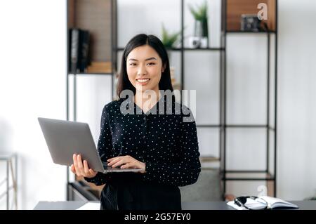 Porträt einer erfolgreichen asiatischen Frau. Eine hübsche, angenehme Japanerin, chinesin oder koreanerin steht im Büro in einem stilvollen formellen Hemd, hält einen offenen Laptop, blickt auf die Kamera und lächelt freundlich Stockfoto