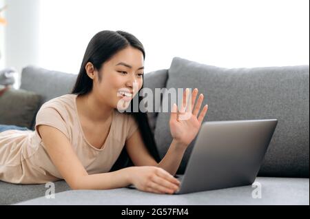 Online-Videokommunikation. Positive niedliche asiatische Mädchen, zu Hause auf einer Couch liegen, nutzt einen Laptop, Gespräche auf einer Videokonferenz mit Freunden oder Familie, chatten in sozialen Netzwerken, begrüßt den Gesprächspartner Stockfoto