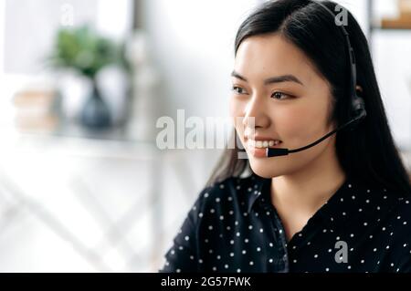 Seitenansicht einer angenehmen, zufriedenen jungen asiatischen Frau mit Headset, Freiberufler, Callcenter-Mitarbeiter oder Berater, Hotline-Betreiber, sieht direkt aus, lächelt freundlich, führt Online-Beratung durch Stockfoto