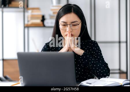 Asiatische junge Frau mit Brille, Büroangestellte, Managerin oder Unternehmerin, die an einem Schreibtisch im Büro sitzt, sich um das Ergebnis sorgt, betet, hofft auf Erfolg, faltete ihre Handflächen nahe ihrem Gesicht Stockfoto