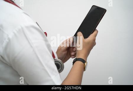 Hände eines Arztes, der das Mobiltelefon im weißen Mantel und das rote Stethoskop auf weißem Hintergrund überprüft. Stockfoto