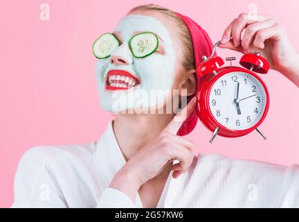 Lächelnde Frau mit kosmetischer Gesichtsmaske und Wecker in der Hand. Kosmetische Verfahren. Beauty Spa und Kosmetik. Stockfoto