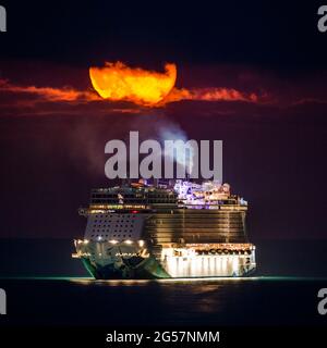 Weymouth, Dorset, Großbritannien. Juni 2021. Wetter in Großbritannien. Der fast volle Erdbeer-Supermond leuchtet orange, während er durch einen kleinen Wolkenbruch über dem leeren NCL-Kreuzschiff Norwegian Escape, das in der Bucht von Weymouth in Dorset vor Anker liegt, leuchtet. Bildnachweis: Graham Hunt/Alamy Live News Stockfoto