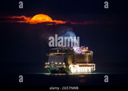 Weymouth, Dorset, Großbritannien. Juni 2021. Wetter in Großbritannien. Der fast volle Erdbeer-Supermond leuchtet orange, während er durch einen kleinen Wolkenbruch über dem leeren NCL-Kreuzschiff Norwegian Escape, das in der Bucht von Weymouth in Dorset vor Anker liegt, leuchtet. Bildnachweis: Graham Hunt/Alamy Live News Stockfoto