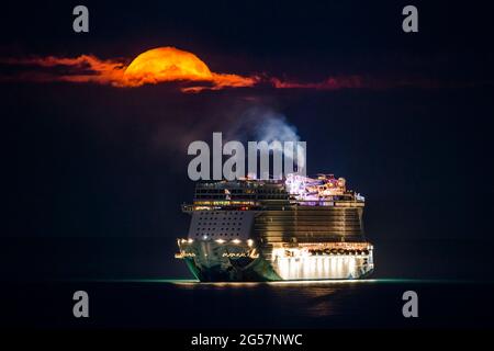 Weymouth, Dorset, Großbritannien. Juni 2021. Wetter in Großbritannien. Der fast volle Erdbeer-Supermond leuchtet orange, während er durch einen kleinen Wolkenbruch über dem leeren NCL-Kreuzschiff Norwegian Escape, das in der Bucht von Weymouth in Dorset vor Anker liegt, leuchtet. Bildnachweis: Graham Hunt/Alamy Live News Stockfoto