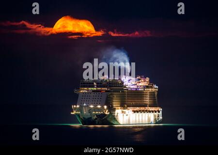 Weymouth, Dorset, Großbritannien. Juni 2021. Wetter in Großbritannien. Der fast volle Erdbeer-Supermond leuchtet orange, während er durch einen kleinen Wolkenbruch über dem leeren NCL-Kreuzschiff Norwegian Escape, das in der Bucht von Weymouth in Dorset vor Anker liegt, leuchtet. Bildnachweis: Graham Hunt/Alamy Live News Stockfoto