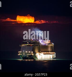 Weymouth, Dorset, Großbritannien. Juni 2021. Wetter in Großbritannien. Der fast volle Erdbeer-Supermond leuchtet orange, während er durch einen kleinen Wolkenbruch über dem leeren NCL-Kreuzschiff Norwegian Escape, das in der Bucht von Weymouth in Dorset vor Anker liegt, leuchtet. Bildnachweis: Graham Hunt/Alamy Live News Stockfoto