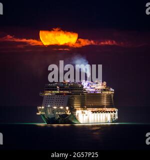 Weymouth, Dorset, Großbritannien. Juni 2021. Wetter in Großbritannien. Der fast volle Erdbeer-Supermond leuchtet orange, während er durch einen kleinen Wolkenbruch über dem leeren NCL-Kreuzschiff Norwegian Escape, das in der Bucht von Weymouth in Dorset vor Anker liegt, leuchtet. Bildnachweis: Graham Hunt/Alamy Live News Stockfoto