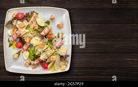 Caesar-Salat mit Kräutern, Hühnerspeck, Eiern und Tomaten auf einem quadratischen hellen Teller, auf dunklem Hintergrund Stockfoto