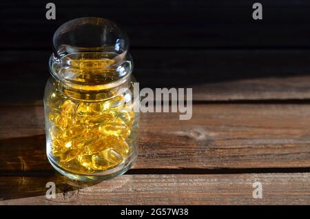 Glas mit Nahrungsergänzungsmitteln in durchsichtigen gelben Kapseln, die vom Sonnenlicht auf einem alten Holztisch beleuchtet werden. Vitamine für Immunität und Langlebigkeit. Stockfoto