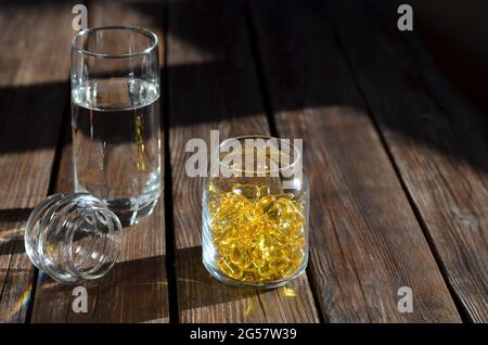 Auf dem Tisch befindet sich ein Glas mit Vitaminen in gelben Gelatinekapseln und einem Glas Wasser, das vom Sonnenlicht beleuchtet wird. Nahrungsergänzungsmittel für gesunde und Stockfoto