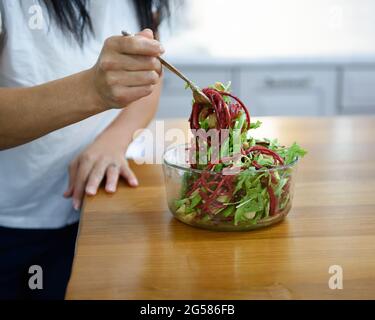 Frauenhände mit einer Gabel, die einen Spinat- und Rübensalat aus einer Glasschüssel auf einer Holztheke verzehrt. Stockfoto